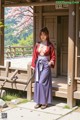 A woman in traditional attire stands outside a wooden structure with cherry blossoms and greenery in the background.